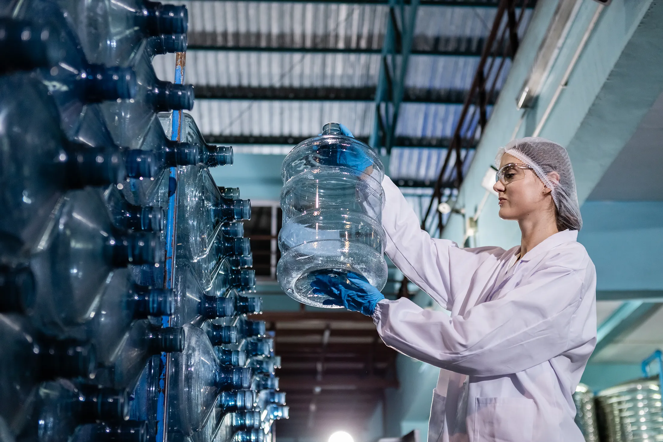 female-worker-holding-checking-quality-of-large-dr-2025-03-10-00-32-20-utc (1) Female worker in protective gear inspecting large water bottles at a bottling facility.