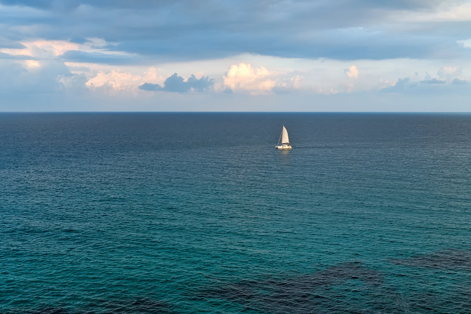 A lone sailboat cruising across a calm open sea under a partly cloudy sky, symbolizing freedom, long-distance sailing, and off-grid marine living.